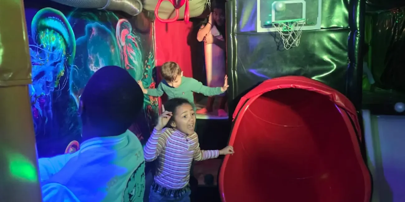 Children playing in the slide play area inside the Essex Soft Play Bus with climbing steps and mini basketball hoop.