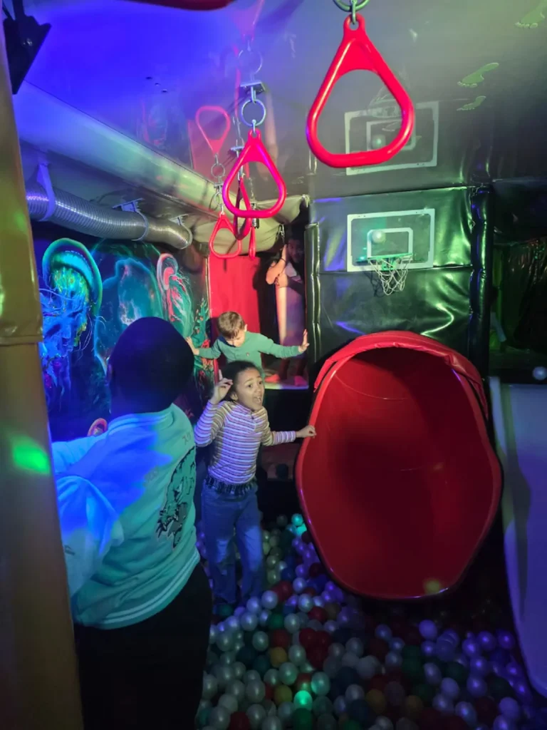 Children playing in the slide play area inside the Essex Soft Play Bus with climbing steps and mini basketball hoop.