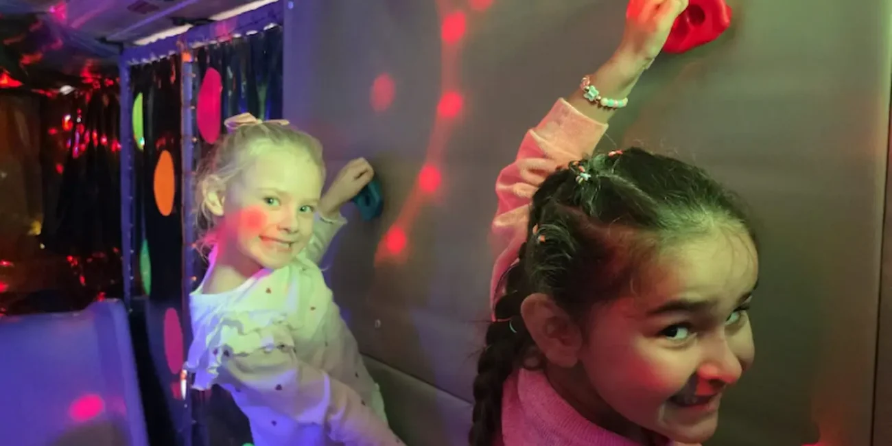 Children using the soft play climbing wall inside the Essex Soft Play Bus with colourful lighting and indoor play equipment.